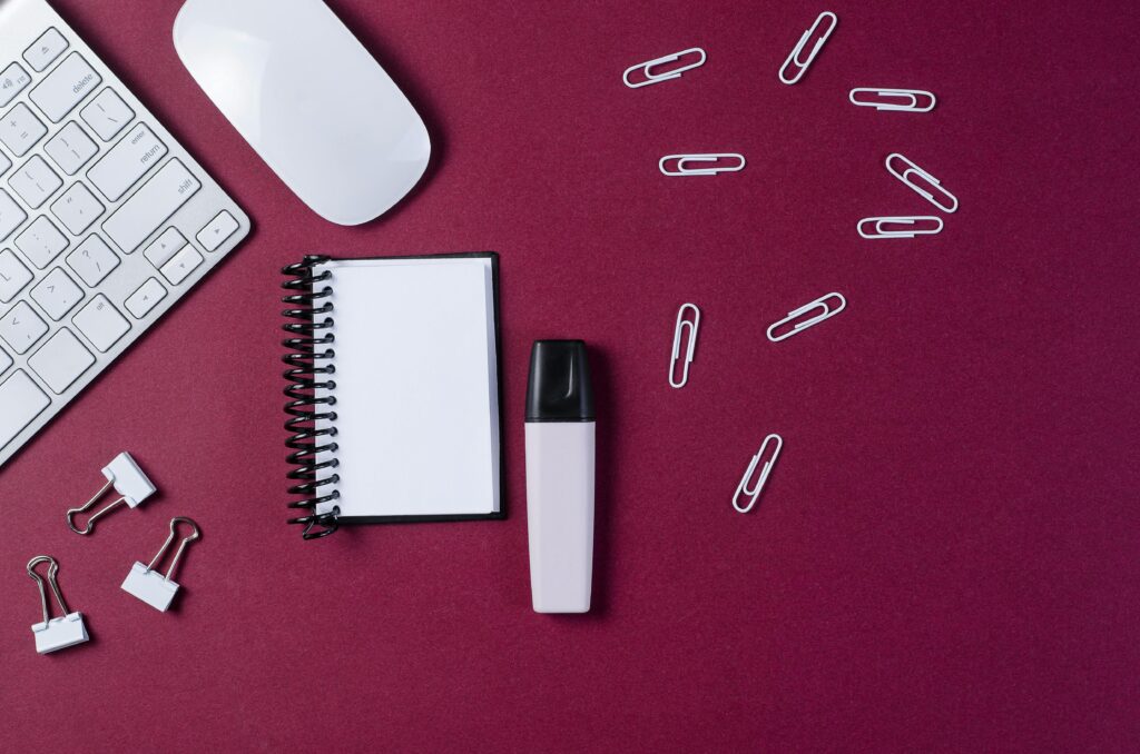 Office stationery on a burgundy desk with copyspace, featuring keyboard, notebook, and paper clips.